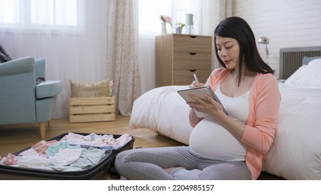 Pretty Asian Mother In Her Last Trimester Is Tucking Her Hair While Counting The Baby Supplies In The Suitcase And Adding Items To Her Shopping List.