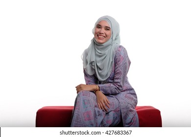 Pretty asian muslim woman wearing traditional malay costume known as songket is smiling while sitting on red sofa on white background
