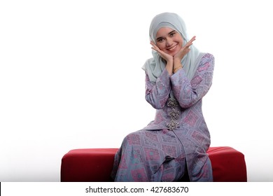 Pretty Asian Muslim Woman Wearing Traditional Malay Costume Known As Songket Is Smiling And Making Pretty Face While Sitting On Red Sofa On White Background
