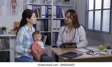 Pretty Asian Pediatrician Answering The Mother’s Questions Patiently In Hospital Room. Korean Parent Holding Child Listening Attentively To The Doctor During The Appointment. Real Moments