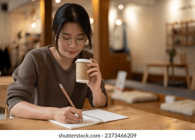 A pretty Asian woman focuses on keeping her diary or making lists in a book and sipping coffee while sitting at a table in a beautiful coffee shop. - Powered by Shutterstock