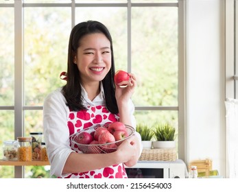Pretty Asian woman holding basket filled with red apples on one hand and hold red apple in the other hand in the kitchen. Healthy eating concept.
