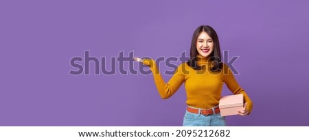 Pretty Asian woman holding gift box and with another hand open to empty space on isolated banner purple background.