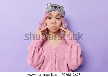 Pretty Asian woman holds breath blows cheeks points with index fingers at face has widely opened eyes wears sleepmask and pink jumper isolated over purple background. Face expressions concept