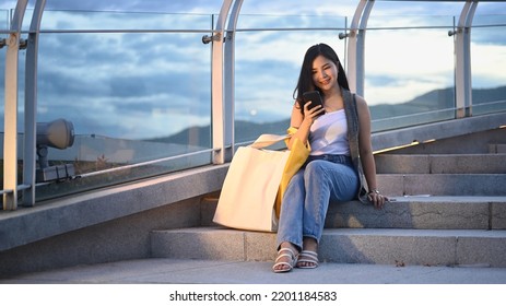 Pretty Asian Woman Sitting Using Mobile Phone And Sitting On Stairs At Terrace With Beautiful Evening Sky In Background