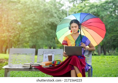 pretty asian woman transgender wears rainbow blouse,raise rainbow umbrella with rainbow handbag sitting and studying with laptop in the park.concept of LGBTQ life, lifestyle,working,studying etc