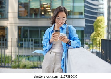 Pretty Asian woman watches video in social networks on smartphone drinks takeaway coffee sends text messages wears spectacles and casual clothes poses against urban building. Modern lifestyle - Powered by Shutterstock