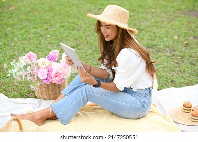 Pretty Asian Young Woman In A Casual Outfit And Picnic Summer Hat Using Portable Tablet During Enjoy Picnic In A Nice Summer Weekend.