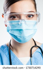 Pretty Attractive Female Doctor,closeup Headshot Portrait,wearing Blue Uniform,safety Goggles,protective Surgical Face Mask,stethoscope. Studio Portrait Isolated On White Background,neutral Expression