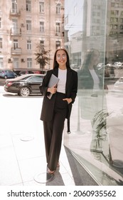 Pretty Attractive Stylish Smiling Young Asian Woman Walks Along A City Street In Black Suit, Spring Fashion, Holds Laptop In Her Hands, Leaning Elbows On Shop Window. Urban Life Concept