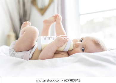 Довольно девочка пьет воду из бутылки, лежащей на кровати. Ребенок носил подгузник в детской комнате.