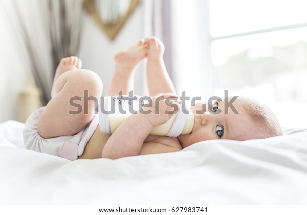 A Pretty baby girl drinks\
water from bottle lying on bed. Child weared diaper in nursery\
room.
