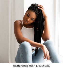 Pretty beautiful afro woman with cool dreadlocks in sexy white fashion top in stylish jeans sits at window near white wall in studio. Attractive African American girl posing in room. Casual style