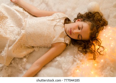 Pretty Beautiful Child Girl Sleep On Fake Snow On The Floor. Fall Asleep After Christmas Party. Little Brunette In White Dress And Curly Hair Laying With Eyes Closed.  