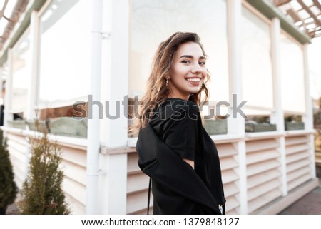 Pretty beautiful young woman with a cute attractive smile in a black T-shirt in a fashionable black coat posing standing on the street near a wooden building. Positive happy girl enjoys spring weekend