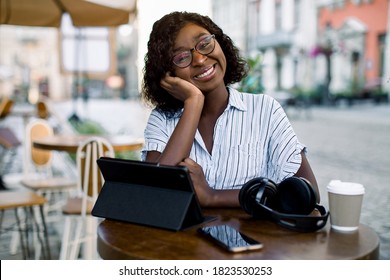 Pretty Black Business Lady , Sitting At The Table In Outdoor City Coffee Shop, With Headphones, Smartphone And Tablet Ipad, Enjoying Her Lunch Time Outside Office, Smiling To Camera