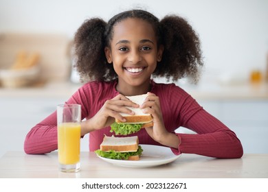 Pretty Black Girl With Bushy Curly Hair Having Snack At Home Alone, Eating Vegetable And Greens Sandwich, Drinking Fresh Orange Juice, Closeup Portrait. Healthy Lifestyle, Diet For Teenagers, Kids