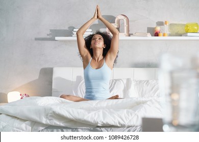 Pretty Black Girl With Curly Hair Sitting In Bed In The Morning. Beautiful Young African American Woman Doing Yoga After Good Sleep.