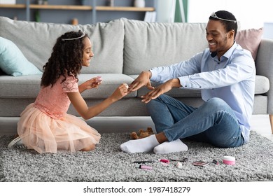 Pretty black girl daughter making manicure for her father, sitting on floor in living room, playing with kids toys. Beautiful african american kid putting nail polisher on her handsome daddy nails