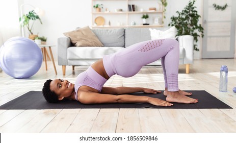 Pretty black lady doing yoga on sports mat, standing in half bridge pose at home. Beautiful African American woman doing asana for strength of abdomen muscles, staying fit and healthy during isolation