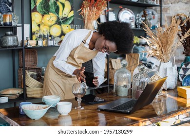 Pretty Black Woman In Apron Creating Online Catalogue Using Wireless Laptop. African Saleswoman In Eyeglasses Taking Picture Of Wine Glass In Shop On Modern Smartphone.