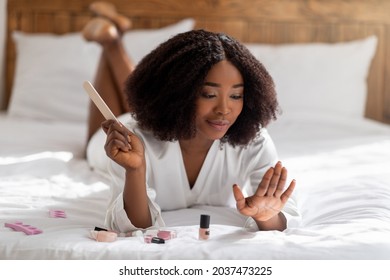 Pretty black woman lying on bed with nail file, doing manicure, taking care of her hands at home. Charming African American lady making self-care procedure indoors. Wellness routines concept