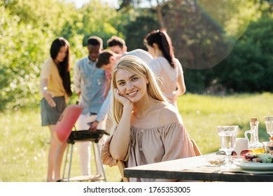Hübsches blondes Mädchen mit Zahnlächeln sitzend auf Stuhl mit Tisch auf interkulturellen Freunden, Grill kochen