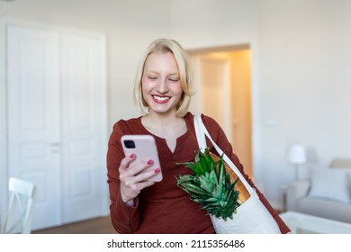 Pretty Blond Woman Arriving Home Carrying Groceries In A Eco Friendly Tote Bag Using Her Smart Mobile Phone - Multi Tasking