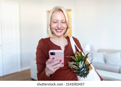 Pretty Blond Woman Arriving Home Carrying Groceries In A Eco Friendly Tote Bag Using Her Smart Mobile Phone - Multi Tasking