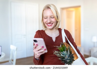 Pretty Blond Woman Arriving Home Carrying Groceries In A Eco Friendly Tote Bag Using Her Smart Mobile Phone - Multi Tasking