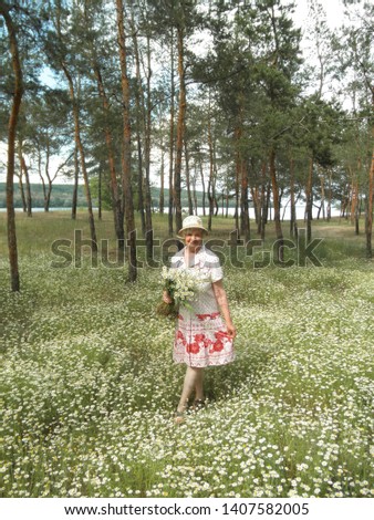 Pretty blond woman in a hat and a white dress with a pink pattern with a bouquet of field daisies posing on a chamomile meadow in a pine forest