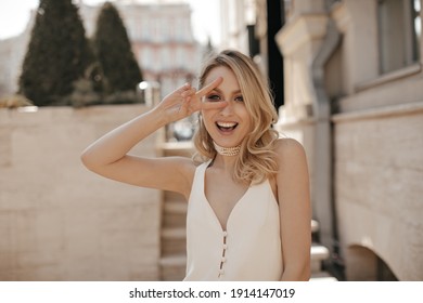 Pretty blonde attractive woman in pearl necklace and white silk dress shows peace sign, looks into camera and smiles outside.