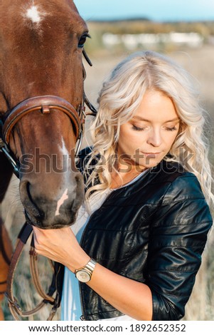 A pretty blonde in a black jacket stands in a field next to a horse at sunset. Love and friendship concept.