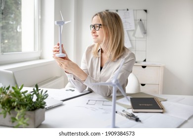 Pretty Blonde Business Woman Is Sitting In Her Office Working On New Alternative Energy Development Looking At A Model Of A Wind Turbine And Is Happy