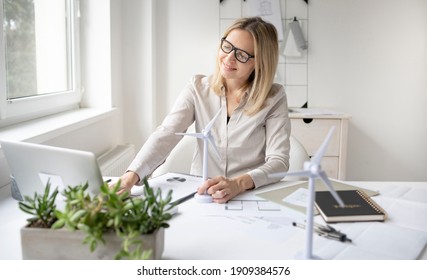 pretty blonde business woman is sitting in her office working on new alternative energy development looking at a model of a wind turbine and is happy