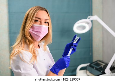 Pretty Blonde Cosmetology Doctor In Pink Disposable Face Mask And Blue Rubber Gloves Sit At Medical Cabinet, Beauty Salon And Fill Syringe With Drug, Filler, Botox For Skin Treatment