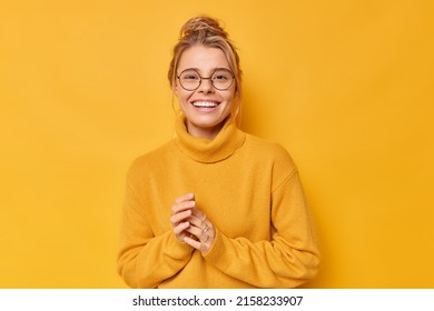 Pretty blonde European woman smiles toothily keeps hands together feels satisfied wears round spectacles and casual loose jumper isolated over vivid yellow background. Happy emotions concept - Shutterstock ID 2158233907