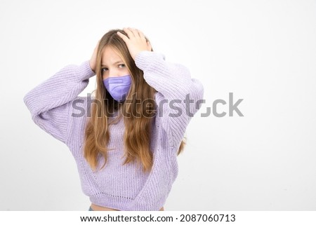 pretty blonde girl in purple wool sweater with nose mouth mask is standing in front of white background