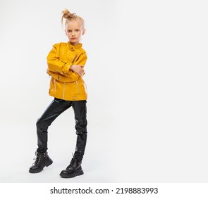 Pretty Blonde Little Girl In A Leather Mustard Jacket, Black Snake Texture Leggings And Boots. High Fashion, Full Length Standing Pose, Isolated Against A Studio Background