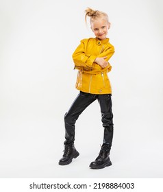 Pretty Blonde Little Girl In A Leather Mustard Jacket, Black Snake Texture Leggings And Boots. High Fashion, Full Length Standing Pose, Isolated Against A Studio Background