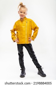 Pretty Blonde Little Girl In A Leather Mustard Jacket, Black Snake Texture Leggings And Boots. High Fashion, Full Length Standing Pose, Isolated Against A Studio Background