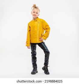 Pretty Blonde Little Girl In A Leather Mustard Jacket, Black Snake Texture Leggings And Boots. High Fashion, Full Length Standing Pose, Isolated Against A Studio Background