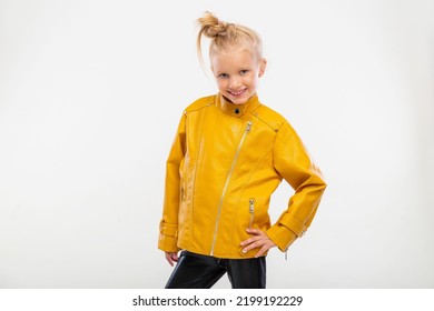 Pretty Blonde Little Girl In A Leather Mustard Jacket, Black Snake Texture Leggings And Boots. High Fashion, Standing Pose, Isolated Against A Studio Background