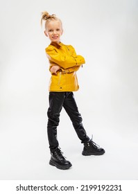 Pretty Blonde Little Girl In A Leather Mustard Jacket, Black Snake Texture Leggings And Boots. High Fashion, Full Length Standing Pose, Isolated Against A Studio Background