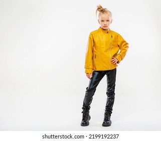 Pretty Blonde Little Girl In A Leather Mustard Jacket, Black Snake Texture Leggings And Boots. High Fashion, Full Length Standing Pose, Isolated Against A Studio Background