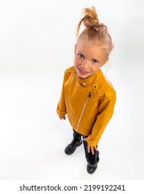 Pretty Blonde Little Girl In A Leather Mustard Jacket, Black Snake Texture Leggings And Boots. High Fashion, Full Length Standing Pose, Isolated Against A Studio Background