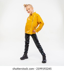 Pretty Blonde Little Girl In A Leather Mustard Jacket, Black Snake Texture Leggings And Boots. High Fashion, Full Length Standing Pose, Isolated Against A Studio Background