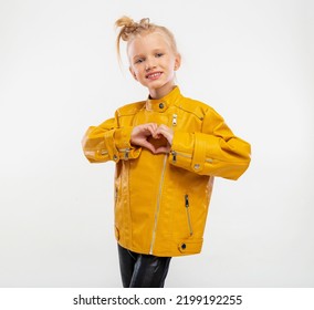 Pretty Blonde Little Girl Shows Heart With Hands In A Leather Mustard Jacket, Black Snake Texture Leggings And Boots. High Fashion, Full Length Standing Pose, Isolated Against A Studio Background