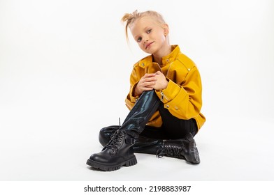 Pretty Blonde Little Girl Sitting In Studio Floor In A Leather Mustard Jacket, Black Snake Texture Leggings And Boots. High Fashion, Full Length Standing Pose, Isolated Against A Studio Background