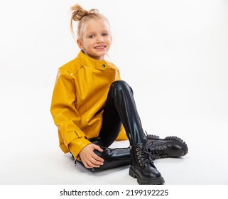 Pretty Blonde Little Girl Sitting In Studio Floor In A Leather Mustard Jacket, Black Snake Texture Leggings And Boots. High Fashion, Full Length Standing Pose, Isolated Against A Studio Background